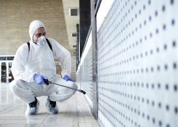Person in white chemical protection suit doing disinfection of public areas to stop spreading highly contagious corona virus.